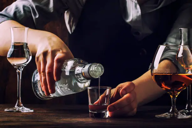 The bartender pours the vodka or tequila in small shot glass on the old bar counter. Vintage wooden background in pub or bar, night mood. Place for text, toning, selective focus