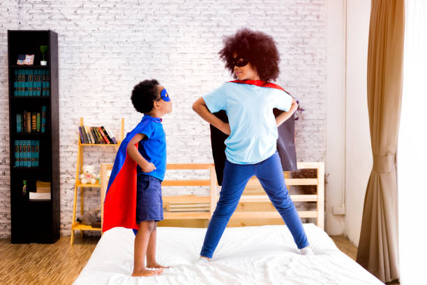 афроамериканец счастливый и уверенный в себе маленькие дети играют и одеваются как супергерой вместе в спальне. - bedroom authority indoors home interior стоковые фото и изображения