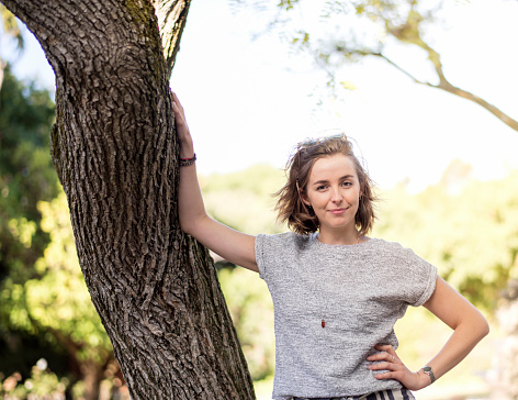 Portrait of smiling woman with hand on hip leaning on tree. Beautiful female is in yard during summer. She is wearing casuals.