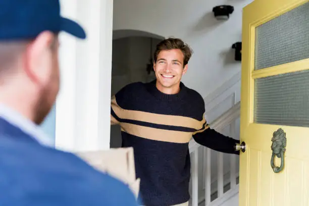 Rear view of worker delivering package to happy male at home. Smiling man looking at delivery person at doorway. He is wearing sweater.