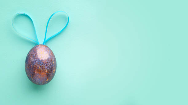 un huevo de pascua en un fondo azul turquesa, con orejas de conejo. concepto de minimalismo - easter text single word paint fotografías e imágenes de stock