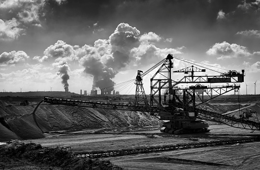 Black and white photograph of an excavator in an open cast mining, a brown-coal burning power plant with pollution and wind turbines in the background.