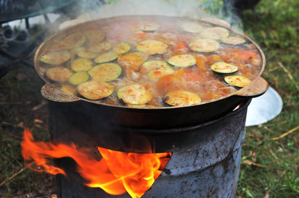 Cooking a hot vegetable dish in brazier in the woods at the stake, close-up; Tourist iron pot with seethe food over the fire in a campsite, bubbles boiling liquid and steam. Summer hike, vacation. cocinar un plato de verduras calientes en brasero en el bosque en la estaca, primer plano; pote turístico del hierro con el alimento del ves sobre el fuego en un camping, burbujas que hierven el líquido y el vapor. caminata de verano, vacaciones.