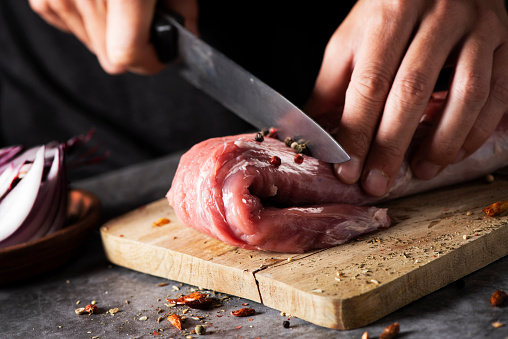 closeup of a young caucasian man cutting a piece of raw pork tenderloin with a knife, on a wooden chopping board, placed on a rustic wooden table or countertop