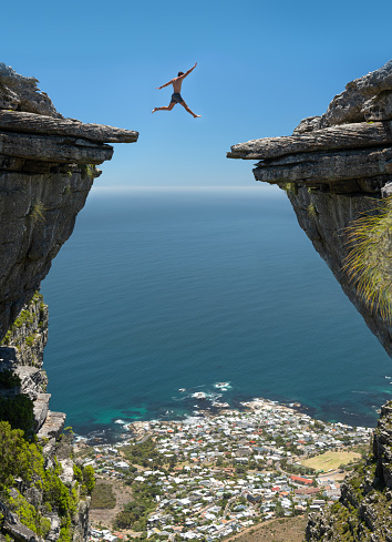 Athletic man jumping the gap between two rocks over a 1000m high cliff with Camps Bay, Cape Town below. Nikon D850. Converted from RAW.