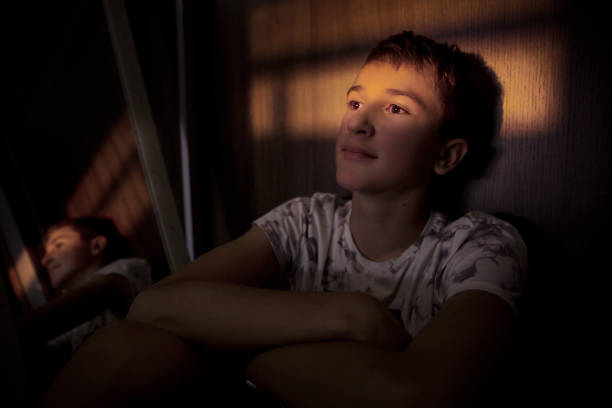 Teenager boy sitting in a dark room beside a mirror, with a light beam over his eyes