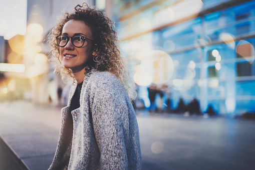Smiling woman in stylish clothing wearing eye glasses outside in the european night city. Bokeh and flares effect on blurred background