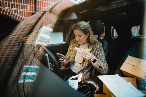 A side-view shot of a mature business woman sitting in the front seat of her car, she is using her mobile phone while she eats her lunch.