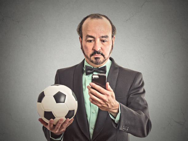 サッカーを保持しているスマート フォンでフットボールの試合を見ているビジネス男 - business team corporate business business person ストックフォトと画像
