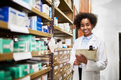 Confident african female pharmacist standing by a medicine shelf holding a clipboard. Druggist at medicine storage room at pharmacy.