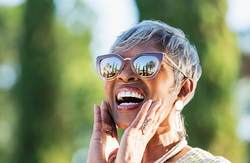 A beautiful senior African-American woman in her 70s wearing sunglasses on a sunny day. She is looking up at the sky, amazed by what she sees, hands touching her chin. A building, palm trees and clear blue skies are visible in the reflection in the sunglasses.