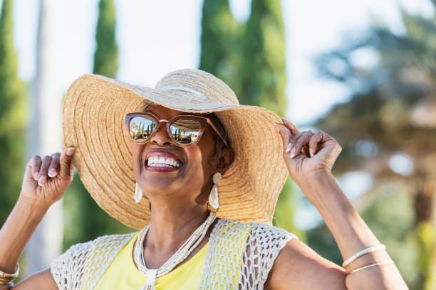 Senior African-American woman wearing sunglasses A beautiful senior African-American woman in her 70s wearing sunglasses and a wide brim hat on a sunny day. She is looking up at the sky, smiling. A building, palm trees and clear blue skies are visible in the reflection in the sunglasses. active seniors summer stock pictures, royalty-free photos & images