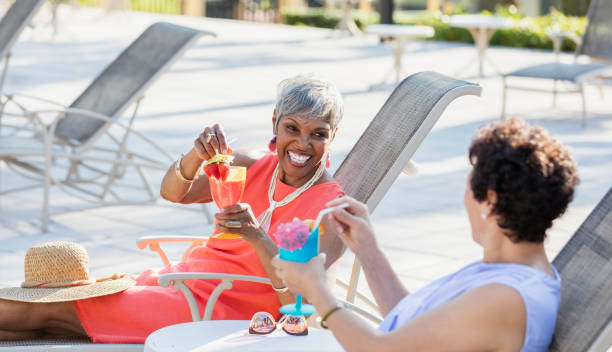 Multi-ethnic senior women by pool with tropical drinks Two multi-ethnic senior women relaxing together on lounge chairs by the pool, having tropical drinks, talking and laughing. The focus is on the African-American woman who is in her 70s. drinks on the deck stock pictures, royalty-free photos & images