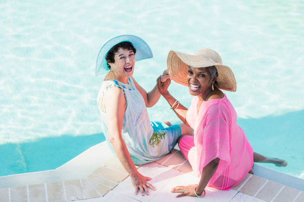Two senior women sitting on pool deck, feet in water