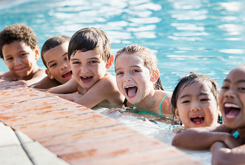 A multi-ethnic group of six children, 6 to 9 years old, having fun at a swimming pool, in the water hanging onto the side, in a row, laughing and looking at the camera.