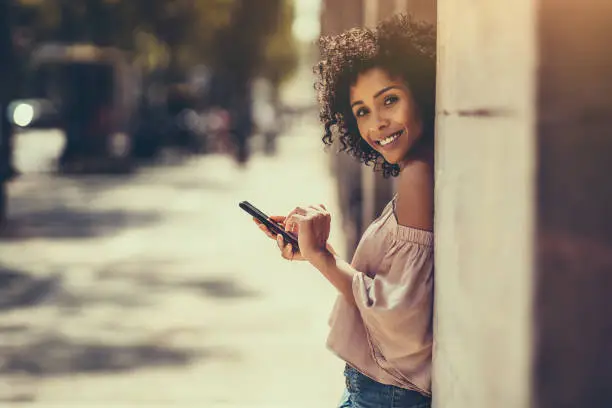 A young cheerful flirtatious curly-hair biracial female outdoors is leaning against the stone column or wall while typing a message using the smartphone, with a copy space place for an advert message