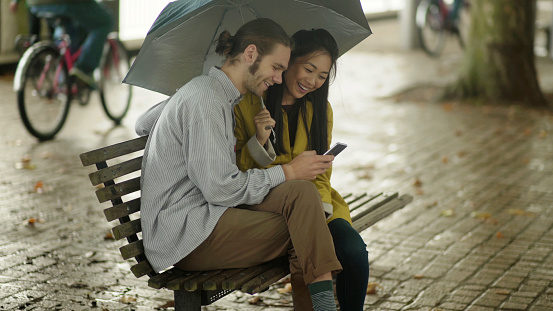 Stock image of a young heterosexual couple (one Caucasian the other Chinese Asian) sitting on an urban bench in the rain, They share a moment with a smartphone in a public space