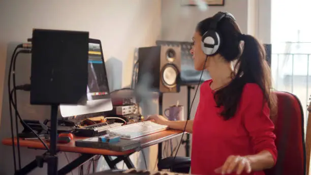 A good looking Hispanic woman is composing music in her private home studio. She is using a computer & music keyboard to compose and is surrounded by musicians’ equipment.