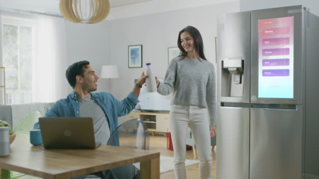 Beautiful Young Woman Opens the Fridge and Gives a Milk Bottle to Her Boyfriend. Then She Checks the Futuristic Digital To-Do List on the Smart Fridge Door.