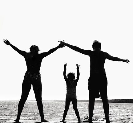 Silhouette of three children, siblings(one teenage boy and one girl, and a four year old girl). Standing, on a picnic table overlooking Lake Michigan in swimwear and holding arms up in enjoyment . Photograph taken in Gladstone, Michigan in The Upper Peninsula. Black and white with high contrast.
