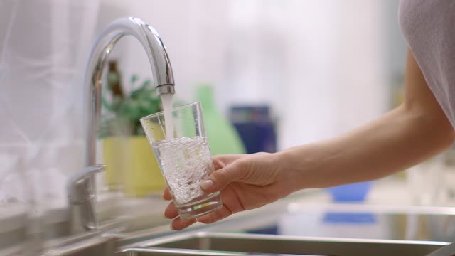 Young woman fills glass under running tap in kitchen.