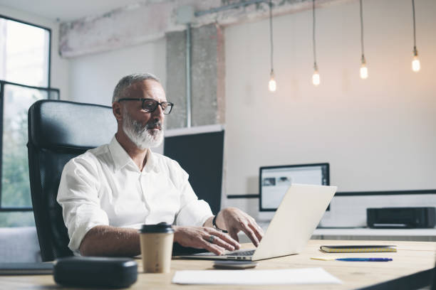 istock Attractive and positive adult businessman using mobile laptop computer while working at the wooden table at modern coworking office. 1128279145