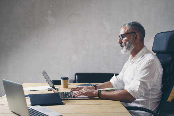 istock Positive Adult businessman using mobile laptop computer while sitting at wooden table at modern coworking place. . 1128280109