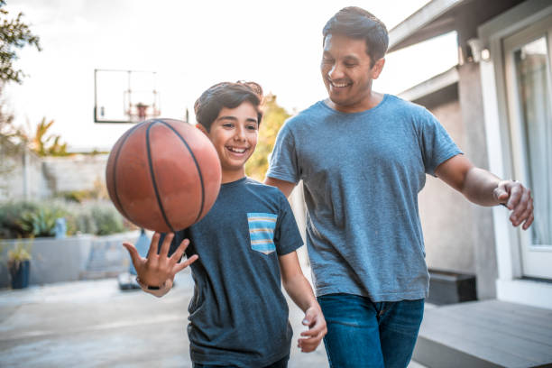 Boy spinning basketball while walking by father Happy boy spinning basketball while walking by father. Mid adult man and child are smiling in backyard. They are wearing casuals during weekend. hispanic stock pictures, royalty-free photos & images