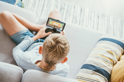 Boy sits on sofa plays with smartphone and gamepad camera top view. Child and electronic devices concept