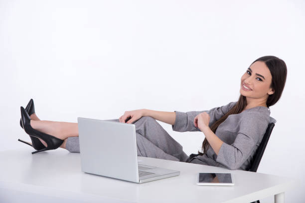 Business woman at her office. She has long black hair and wearing a grey jumpsuit. She has her legs on the table and looking at the camera.