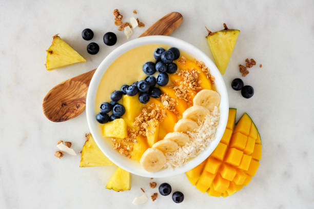 Pineapple, mango smoothie bowl with coconut, bananas, blueberries and granola, above view on a bright background Healthy pineapple, mango smoothie bowl with coconut, bananas, blueberries and granola. Above view scene on a bright background. smoothie stock pictures, royalty-free photos & images