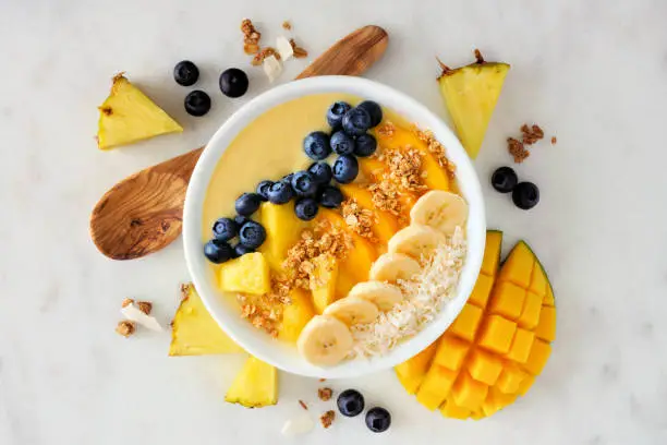Healthy pineapple, mango smoothie bowl with coconut, bananas, blueberries and granola. Above view scene on a bright background.