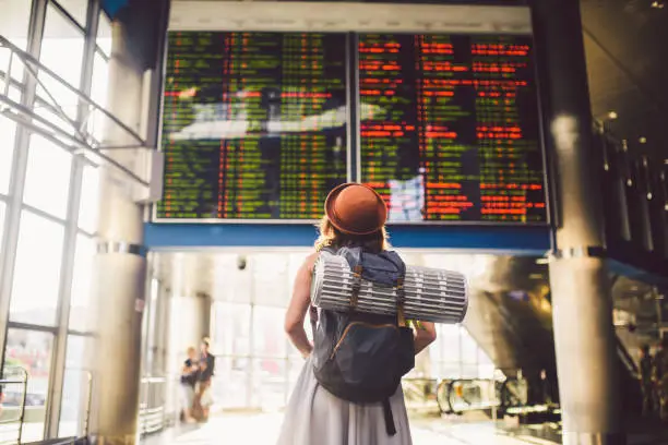 Photo of Theme travel public transport. young woman standing with back in dress and hat behind backpack and camping equipment for sleeping, insulating mat looks schedule on scoreboard airport station