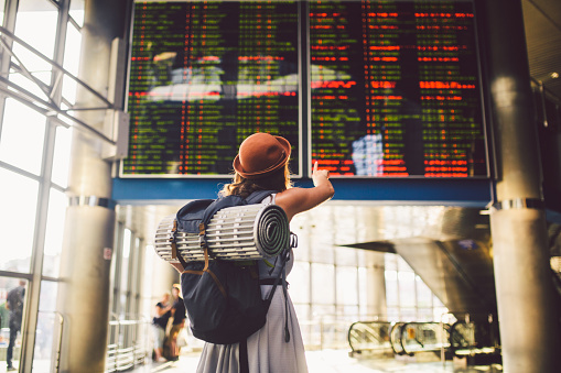 Theme travel public transport. young woman standing back in dress and hat behind backpack camping equipment for sleeping, insulating mat looks schedule on scoreboard airport station hand to the front.