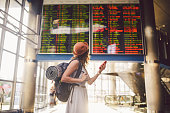 Theme travel and tranosport. Beautiful young caucasian woman in dress and backpack standing inside train station or terminal looking at a schedule holding a red phone, uses communication technology