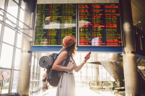 Theme travel and tranosport. Beautiful young caucasian woman in dress and backpack standing inside train station or terminal looking at a schedule holding a red phone, uses communication technology Theme travel and tranosport. Beautiful young caucasian woman in dress and backpack standing inside train station or terminal looking at a schedule holding a red phone, uses communication technology. cancellation photos stock pictures, royalty-free photos & images
