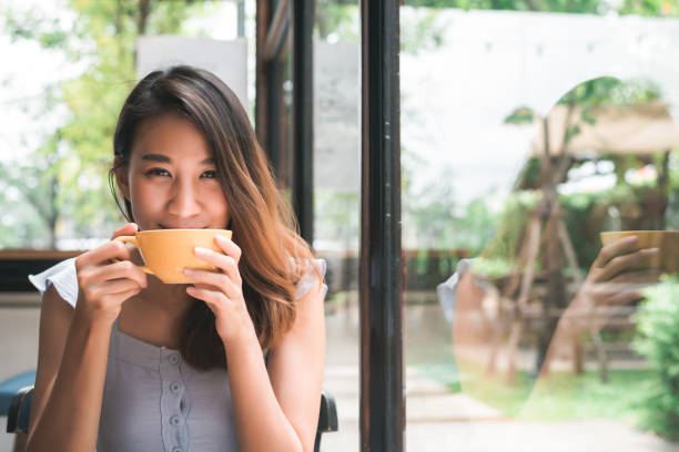 fröhliche asiatische junge frau, die warmen kaffee oder tee trinkt, während sie im café sitzt. attraktive glückliche asiatische frau mit einer tasse kaffee. - tee warmes getränk stock-fotos und bilder