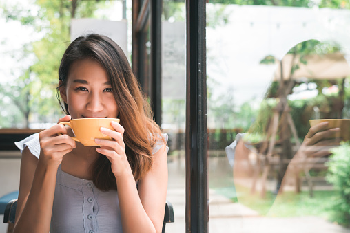 Mujer asiática joven alegre beber café caliente o té disfrutando mientras estaba sentado en la cafetería. Atractiva mujer asiática feliz sosteniendo una taza de café. photo