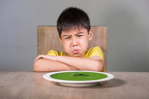 7 or 8 years old upset and disgusted Asian kid sitting on table refusing to eat spinach thick soup looking unhappy rejecting vegetarian food in child hate green vegetables and healthy nutrition stock photo