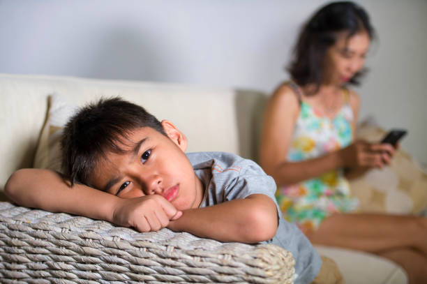 young sad and bored 7 or 8 years old Asian child at home couch feeling frustrated and unattended while mother networking on mobile phone as internet addict neglecting her son stock photo