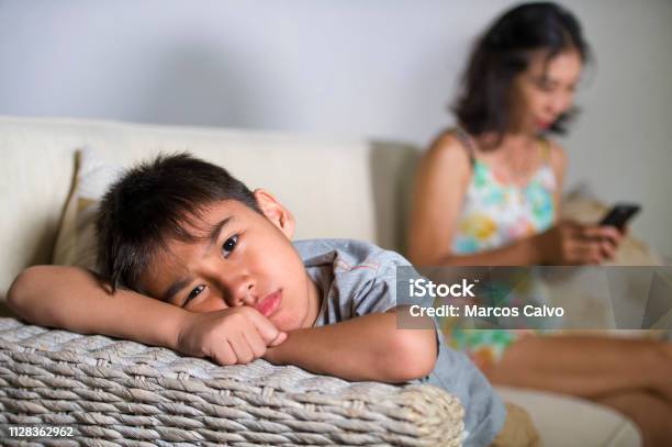 Young Sad And Bored 7 Or 8 Years Old Asian Child At Home Couch Feeling Frustrated And Unattended While Mother Networking On Mobile Phone As Internet Addict Neglecting Her Son Stock Photo - Download Image Now