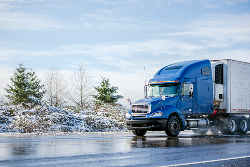 Big rig pro long haul blue semi truck tractor transporting commercial cargo in refrigerator semi trailer going on the wet glossy road with water from melting snow and winter snowy trees on the side