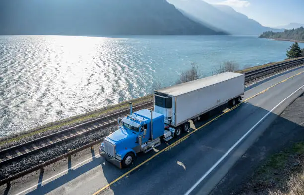Photo of Blue classic American bonnet big rig semi truck trancporting cargo in refrigerated semi trailer moving on the road along the river in Columbia River Gorge area