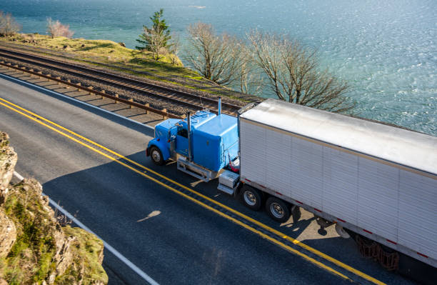Blue classic big rig with reefer semi trailer transporting cargo running on the road along the Columbia River Big rig long haul bonnet American semi truck transporting commercial cargo in refrigerated semi trailer running on the narrow road along the Columbia River and mountain in Columbia River Gorge car carrier semi stock pictures, royalty-free photos & images