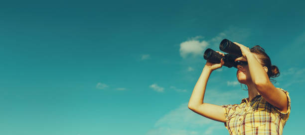 Young scout girl looks through binoculars on blue sky background