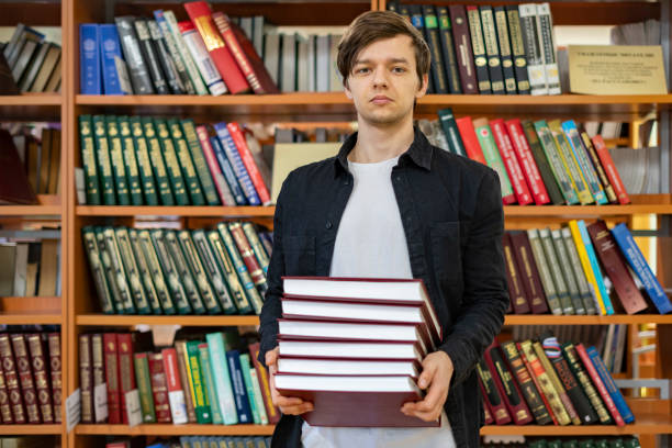 istock man in university holding stack of books (book shelf background) b 1128391473