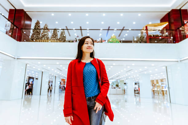 Low angle view of Chinese woman in her 30s wearing red jacket and jeans, walking through shopping mall, smiling, happiness, window shopping