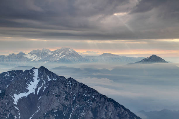 Sun rays burst through dark gray cloudy sky over rocky ridges of Karavanke and Kamnisko Savinjske Alpe ranges in haze, Begunjscica Grintovec Storzic peaks Gorenjska Kranjska Carniola Slovenia Central Europe