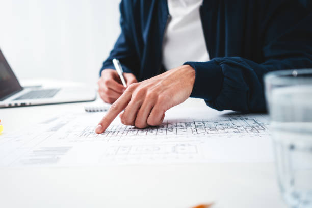 istock Close-up of hands architect uses laptop and building blueprint on working table in office space. Young engineer working on building plan 1128407564