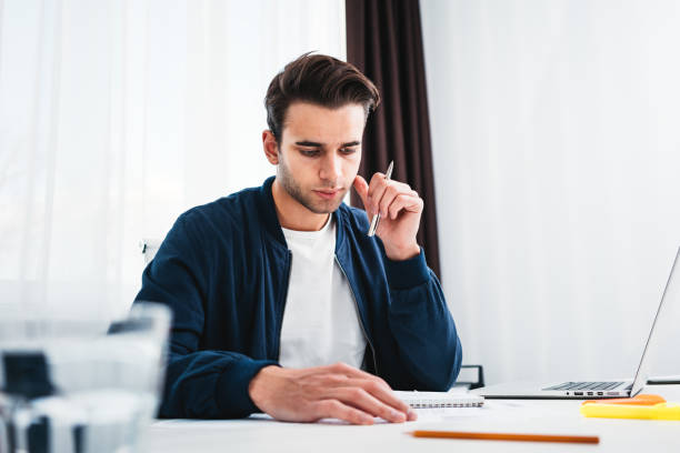 istock Concentrated man writing new ideas in notepad looking on screen laptop. Young coworker sits at table and working in office 1128407579